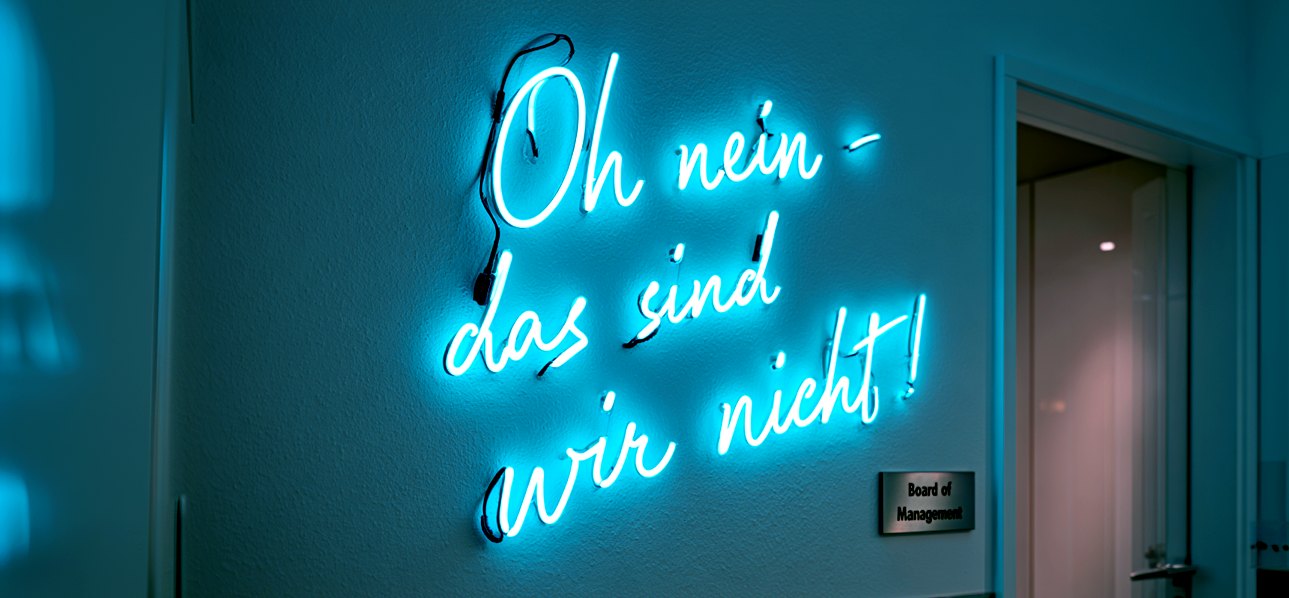 Türkis leuchtende Neonschrift „Oh nein – das sind wir nicht!“ an einer Wand neben einer Tür mit dem Schild „Board of Management“.